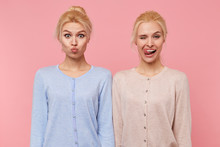 Portrait Of Beautiful Young Blonde Twins Fool Around And Make Faces Isolated Over Pink Background. One Girl Sends A Kiss And The Second One Tongues Tooth And Winks.