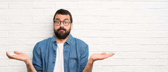 handsome man with beard over white brick wall having doubts while raising hands