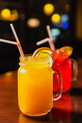 Wall Mural - Two refreshing cold summer lemonades in glass jars, orange and red, served with ice, orange and lime slices. Shot in a bar using studio lights, reflections in a bar stand.