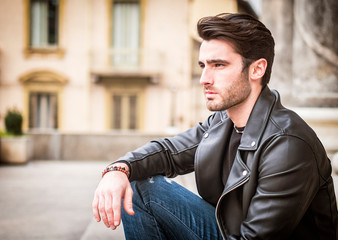 Wall Mural - One handsome young man in urban setting in modern city, standing, wearing black leather jacket and jeans, looking away