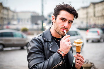 Attractive young man standing, wearing black leather jacket while eating a tasty ice cream outdoors in city