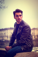 Wall Mural - One handsome young man in urban setting in modern city, standing, wearing black leather jacket and jeans, looking away