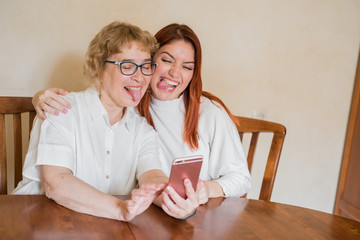 Mom and daughter are photographed on the front camera of the smartphone while sitting at home at the table. Cheerful elderly mother and her daughter making comic selfies and showing tongues.