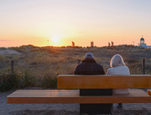 Elderly Couple (woman And Man)is Sitting On The Bench By The Sea At The Sunset In Summer