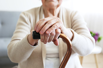 Wall Mural - Elderly woman sitting in nursing home room holding walking quad cane with wrinked hand. Old age senior lady wearing beige cardigan, metal aid stick handle bar close up. Interior background, copy space