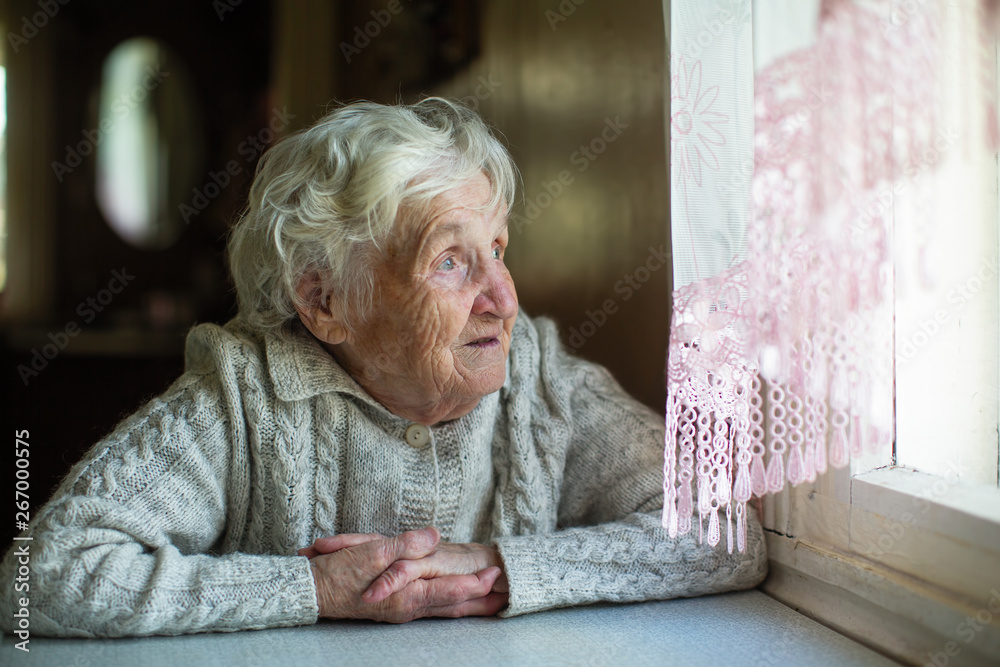 An elderly woman looks out the window sitting in his house. Care of lonely pensioners. - obrazy, fototapety, plakaty 