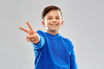 Wall Mural - childhood, gesture and people concept - portrait of smiling little boy in blue hoodie showing peace hand sign over grey background