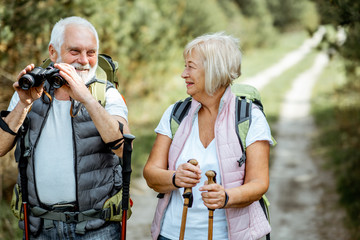 Wall Mural - Portrait of a happy senior couple standing together with binoculars, backpacks and trekking sticks while hiking in the forest. Concept of an active lifestyle on retirement