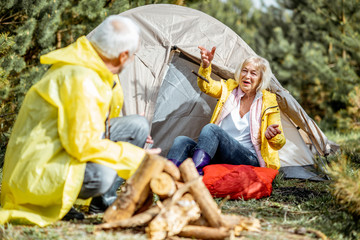 Wall Mural - Senior couple in yellow raincoats having fun while making fireplace at the campsite near the tent in the woods