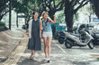© PR Image Factory - two young asian girls walking on city street under trees shadow. full length beautiful female friends travelers relax outdoor excited point finger sharing amazing view in urban travel taiwan taipei