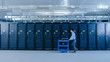 © Gorodenkoff - In the Modern Data Center: IT Technician Working with Server Racks, Pushes Cart Between Rows of Server Racks. On a Pushcart New Hardware for System Update. Engineer Doing Maintenance and Diagnostics.