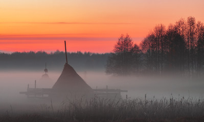 Rural meadow with haystacks in the foggy evening in red-orange light of the sunset sky