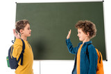 Fototapeta Łazienka - two smiling schoolboys with backpacks showing high five sign near blackboard isolated on white