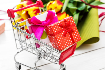 Wall Mural - Shopping cart with gift boxes and bouquet of tulips on white wooden table. close up. Holiday concept.