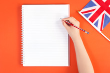 Hands On A Desk With A Pencil And A Blank Notebook And British English Union Jack Flag On Orange Background. Flat Lay With Copy Space. Education Study Or English Language Learning Concept