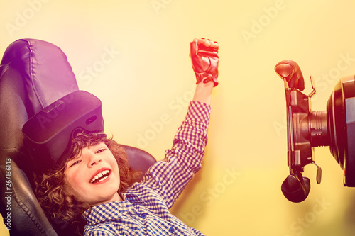 Cuadros en Lienzo Toddler boy with a virtual reality headset sitting in a autosimulator triumphantly raising his hand up on a yellow background