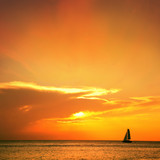 Fototapeta Zachód słońca - Orange color seascape image with shiny sea and sailboat over cloudy sky and sun during sunset in Cozumel, Mexico