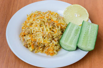 Appetizing, tasty, hot, fresh pilaf (pilau, pilaw, pilaff) on a plate with two cucumbers and a slice of lemon. Pilaf with beef, carrots, onions, garlic on wooden background. A traditional dish Asia.