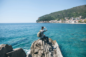 Wall Mural - A tourist with a backpack alone sits on a cliff by the sea and looks into the distance at the view of Petrovac in Montenegro.