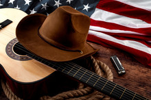 American Culture, Living On A Ranch And Country Muisc Concept Theme With A Cowboy Hat, USA Flag, Acoustic Guitar, Harmonica And A Rope Lasso On A Wooden Background In A Old Saloon