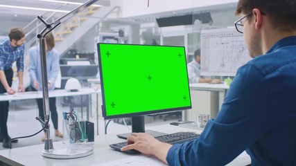 Leinwandbilder - Over the Shoulder Shot of Engineer Working with Green Mock-up Screen Desktop Computer. In the Background Engineering Facility with Specialists Working on Blueprints and Drawings with Industrial Design
