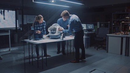 Leinwandbilder - Late at Night in Robotics Engineering Facility Three Technical Engineers Talk and Work on a Wheeled Robot Prototype. In the Background High Tech Research Center with Screens Showing Industrial Design
