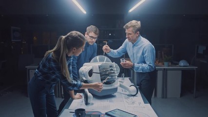 Leinwandbilder - Late at Night in Robotics Engineering Facility Three Technical Engineers Talk and Work on a Wheeled Robot Prototype. In the Background High Tech Research Center with Screens Showing Industrial Design