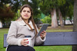 © Mangostar - Pensive Latin girl reading book on tablet screen in park. Young woman in warm jacket sitting on bench, drinking coffee and holding gadget. Outdoor coffee break concept
