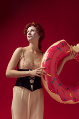 Wall Mural - Medieval redhead young woman as a duchess in black corset and night clothes standing on red background with a swim circle as a donut. Concept of comparison of eras, modernity and renaissance.