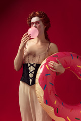 Wall Mural - Medieval redhead young woman as a duchess in black corset and night clothes standing on red background with a swim circle as a donut. Concept of comparison of eras, modernity and renaissance.