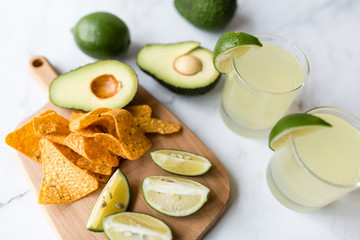 Fresh avocado, lime, drink and nacho chips lying on marble background. Recipe for Cinco de Mayo party