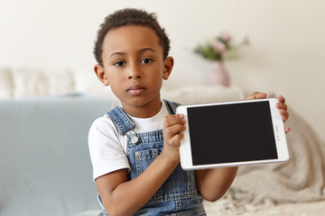 Wall Mural - Happy childhood, ethnicity, rest and relaxation. Happy adorable eight year old African boy lying on stomach on carpet at home, keeping hands on his cheeks, smiling joyfully as if watching cartoons