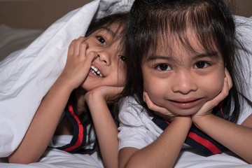  happy little girls twins sister in bed under the blanket having fun, smiling and wacky