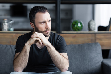 Wall Mural - serious bearded man sitting on sofa with folded hands and looking away