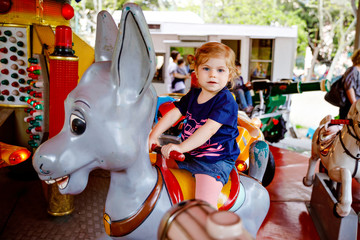 Adorable little toddler girl riding on animal on roundabout carousel in amusement park. Happy healthy baby child having fun outdoors on sunny day. Family weekend or vacations