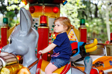 Adorable little toddler girl riding on animal on roundabout carousel in amusement park. Happy healthy baby child having fun outdoors on sunny day. Family weekend or vacations