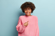 Human reaction concept. Positive dark skinned woman has crisp hair, points at herself, looks gladfully, wears oversized pink jumper, cannot believe she is winner, isolated over blue studio wall