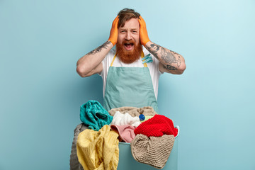 Horrified ginger young man keeps hands on head, opens mouth widely, wears casual blue basin, overstained with housekeeping, stands in front of basin with laundry. Annoyed male householder indoor