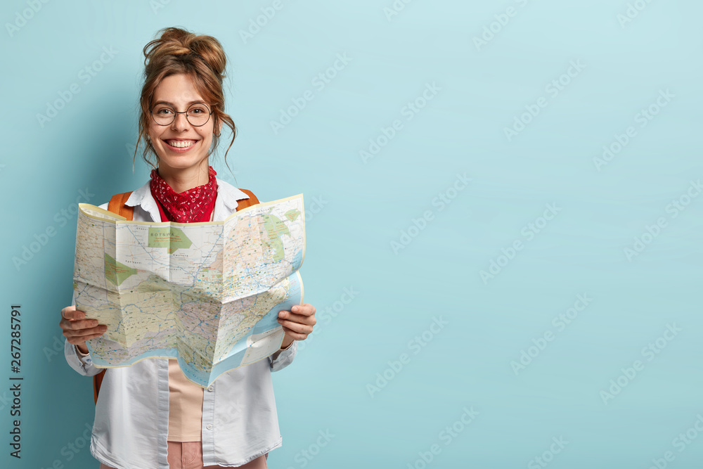 Positive smiling young female tourist searches for inspiring places, holds paper map, finds new sightseeing for discovering, wears round spectacles, red bandana, isolated on blue studio wall - obrazy, fototapety, plakaty 