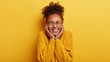 Portrait of cheerful woman feels entertained and amused, watches funny show, touches cheeks with both hands, shows white teeth, keeps eyes closed, wears yellow jumper, stands indoor. Emotions concept