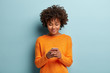 Horizontal shot of beautiful satisfied female teenager focused in smartphone device, chats online with friends, checks notification received on email, being always in touch, wears casual orange jumper