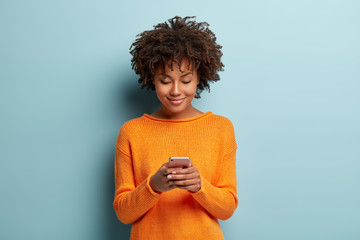horizontal shot of beautiful satisfied female teenager focused in smartphone device, chats online wi