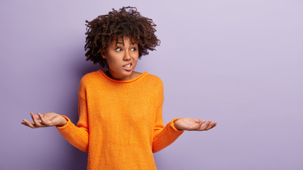Wall Mural - Photo of doubtful Afro woman faces dilemma, spreads hands sideways, clenches teeth, focused aside has difficult decision, dressed casually stands over purple wall with empty space for your information