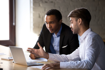 African intern receiving instructions from caucasian colleague sitting using laptop