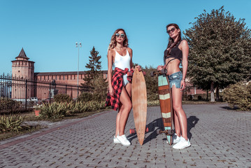 Two girlfriends girls, women sisters, posing in summer in the city, skate board, casual wear shorts swimsuit shirt. The concept of a weekend getaway, fashion style, modern lifestyle. Free space.