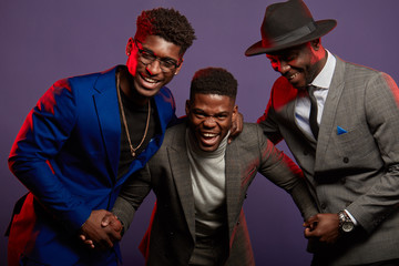 Studio shot of group of African male young adult friends wearing smart suits having fun and laughing over dark violet background