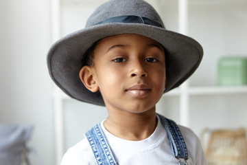 Wall Mural - Style, beauty, children’s wear and fashion concept. Close up picture of adorable fashionable eight year old Afro American boy posing indoors wearing stylish round men's hat and denim jumpsuit