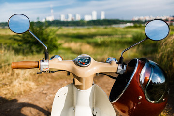 A first-person view of the steering wheel of a classic vintage moped with a beautiful helmet with a mirror visor on it. Ahead is a spacious field of green grass, country road. In the distance you can 