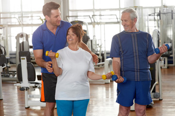 Wall Mural - Handsome male trainer instructing senior woman in gym. Elderly people working out at fitness club. Sport, recreation, healthy lifestyle.