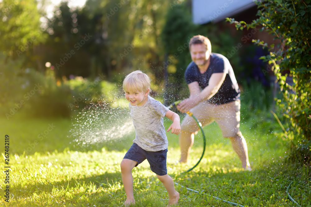Funny little boy with his father playing with garden hose in sunny backyard. Preschooler child having fun with spray of water. - obrazy, fototapety, plakaty 